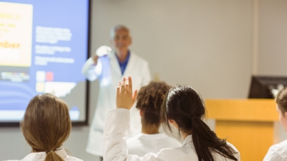 An instructor wearing a white coat teaches at a podium in a medical school classroom.
