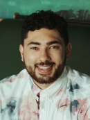 Carlos Storck-Martinez has short black hair and a beard and is wearing a multicolored collared shirt